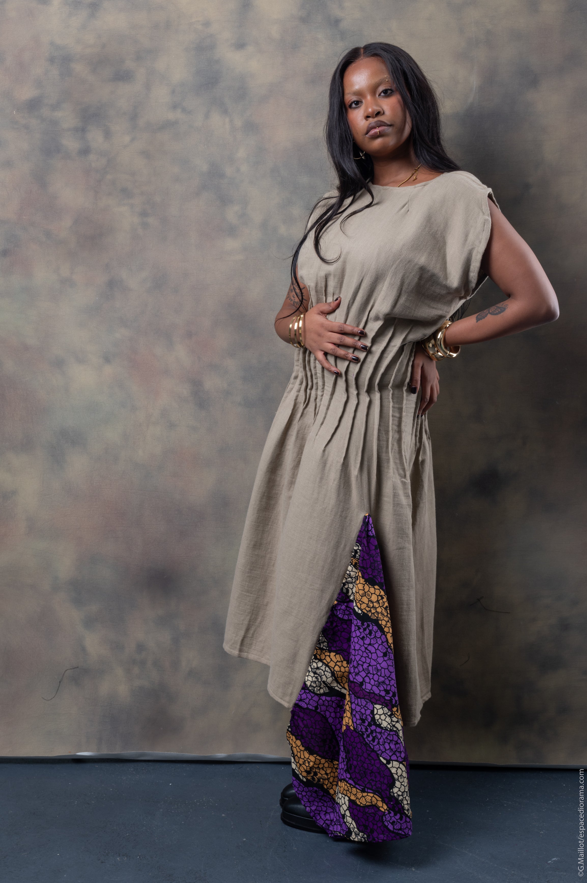 A model wearing a beige linen dress with ruched detailing at the waist and an Ankara fabric panel in purple and gold at the hem.
